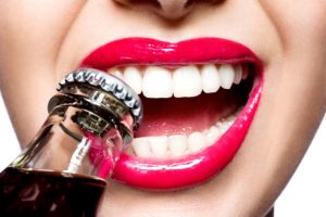 Nose to chin view of a woman with pink lip gloss biting the cap of a soda bottle with her teeth