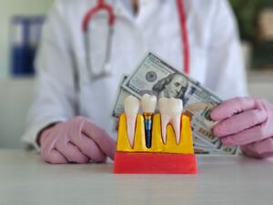 Dentist's gloved hands holding money next to model dental implant